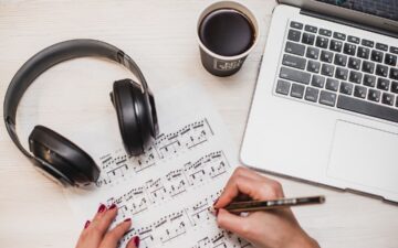headphones and laptop on a desk with sheet music in front and hand that is writing on it.